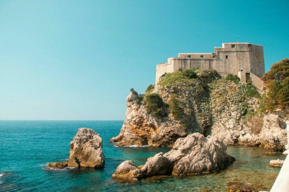 Lovrijenac-fortet i Dubrovnik, Kroatia, ruver majestetisk på en klippe over det krystallklare Adriaterhavet. Fortet, også kjent som «Dubrovniks Gibraltar», er en av byens mest imponerende historiske bygninger og har spilt en viktig rolle i byens forsvar gjennom århundrene. (Foto: Canva) Bildet viser Lovrijenac-fortet i Dubrovnik, Kroatia, som ruver majestetisk på en klippe over det krystallklare Adriaterhavet. Fortet, også kjent som 'Dubrovniks Gibraltar,' er en av byens mest imponerende historiske bygninger og har spilt en viktig rolle i byens forsvar gjennom århundrene. I forgrunnen ser vi store klipper som stikker opp av det turkise vannet, mens den blå himmelen gir en rolig og idyllisk bakgrunn. Grønne vekster klamrer seg til fjellsidene, noe som tilfører et naturlig preg til det robuste landskapet. Dette er en av Dubrovniks mest ikoniske severdigheter, kjent både for sin historie og for sin rolle som innspillingssted i den populære TV-serien Game of Thrones. Perfekt for historieentusiaster og eventyrlystne reisende som ønsker å utforske den unike kombinasjonen av natur og kultur i Kroatia.