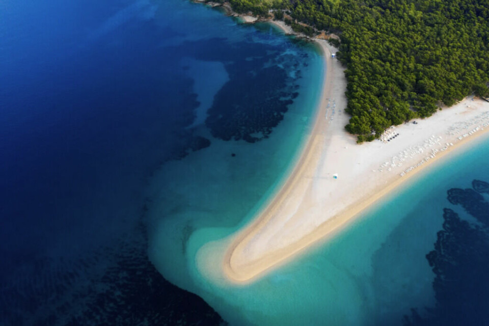 Zlatni Rat, en av Kroatias mest ikoniske strender, som strekker seg ut i det dype blå Adriaterhavet fra øya Brač. (Foto: Canva) Bildet viser en spektakulær luftfoto av Zlatni Rat, en av Kroatias mest ikoniske strender, som strekker seg ut i det dype blå Adriaterhavet fra øya Brač. Den unike, trekantede formen på stranden endrer seg med vind og havstrømmer, noe som gir den et dynamisk og stadig skiftende utseende. Vannet rundt stranden har en nydelig gradient av blåtoner, fra lys turkis nær kysten til dyp marineblå lenger ut. På høyre side ser man frodig grønn vegetasjon, som gir en vakker kontrast til den hvite sanden og det klare havet. Dette er en av de mest populære strendene i Kroatia, kjent for sine fantastiske badeforhold, vannsportmuligheter og naturskjønnhet. Et perfekt reisemål for sol- og strandelskere som ønsker en eksotisk ferie ved Adriaterhavet!