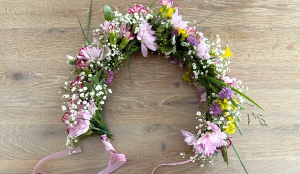 Blomsterkrans med silkebånd – lett å tilpasse ved å knyte den i nakken. Blomsterkrans laget av rosa, lilla og hvite blomster, med silkebånd i endene. Ligger på trebord. Egnet som hårpynt til midtsommer og sommerfest. Kransen er åpen og knytes i nakken.