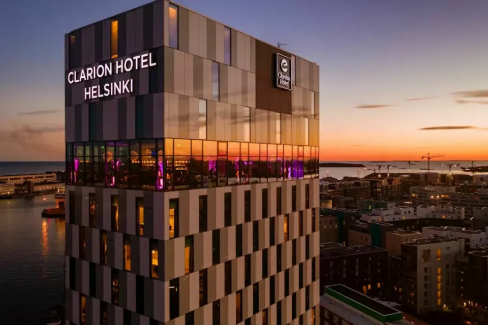 Clarion Hotel® Helsinki – ikonisk designhotell ved sjøen, med spektakulær utsikt over byen og solnedgangen fra øverste etasje. (Foto: Strawberry) Bildet viser Clarion Hotel® Helsinki i solnedgang. Hotellet er en høy og moderne bygning ved vannet, med en fasade i lyse og mørke vertikale paneler som skaper et grafisk mønster. Øverst på bygget, i de øverste etasjene, vises store vindusflater hvor man ser inn i et opplyst område med lilla og gyldne lys – trolig hotellets Skybar eller restaurant. Bygget bærer tydelig skilt med teksten “Clarion Hotel Helsinki” og Clarion-logoen. I bakgrunnen ser man byen strekke seg utover med lavere bygninger, havneområde, kraner og vann som glitrer i solnedgangen. Himmelens farger går fra varm oransje til dyp blå og skaper en rolig og vakker stemning.