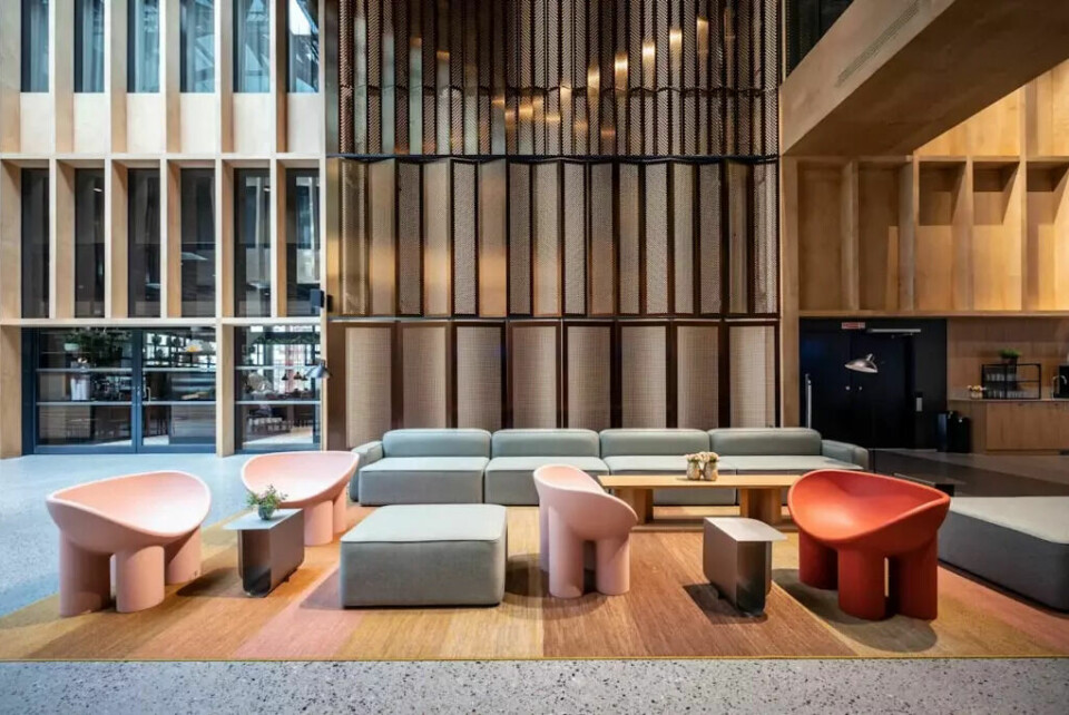 Loungeområdet på Quality Hotel™ Hasle Linie – en stilig og innbydende møteplass med skandinavisk design og moderne komfort. (Foto: Strawberry) Bildet viser en moderne og elegant lounge eller lobbyområde, mest sannsynlig fra Quality Hotel Hasle Linie. Rommet har høyt under taket og er fylt med naturlig lys. Interiøret er preget av skandinavisk minimalisme med lyse trematerialer, perforerte metallpaneler og rene linjer. I forgrunnen står fire lave, runde stoler – to i lys rosa og én i rød – med lekent og skulpturelt design. Disse omkranser et lavt bord i metall og står på et gulvteppe i varme nyanser av rosa, oransje og brun. Ved siden av står en lang, grå modulsofa med myke linjer og lav sittestilling, perfekt for uformelle samtaler eller avslapning. I bakgrunnen sees store vinduer og dører som fører til et annet rom med mer servering eller sosialt område.
