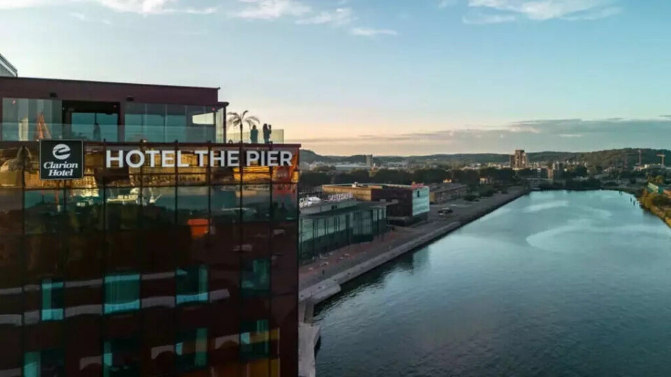 Clarion Hotel® The Pier – et stilig og urbant hotell ved vannet, med takterrasse og utsikt over elven og byen. Perfekt for moderne reisende. (Foto: Strawberry) Bildet viser Clarion Hotel® The Pier, et moderne hotell som ligger helt nede ved vannkanten i en urban del av byen. Fasaden er dekket av mørkt glass som reflekterer havneområdet og bygningene rundt. Øverst på hotellet, ut mot vannet, står det store hvite bokstaver med teksten 'HOTEL THE PIER' og Clarion-logo. På takterrassen ser man flere personer som nyter utsikten, omgitt av glassrekkverk og palmer – noe som gir et eksklusivt og avslappet inntrykk. Til høyre i bildet renner en bred elv eller kanal inn mot land, omgitt av moderne bygg, grønne åser og blå himmel i bakgrunnen.