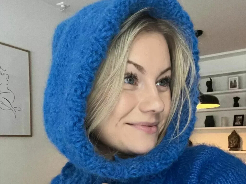 Denne stilige balaklavaen, designet og strikket av Emilie fra Knitting by Emilie, er enkel å lage selv! (Foto: Knitting by Emilie) En kvinne med blondt hår og blå øyne smiler og ser til siden. Hun har på seg en blå, strikket balaklava laget av mykt garn. Bakgrunnen viser en moderne stue med dekorative hyller.