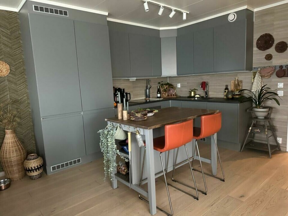Kjøkkenet før oppussingen – Jeg ønsket et varmere uttrykk enn de grå frontene kunne gi. (Foto: Lise Wolden) Et moderne kjøkken med grå fronter, mørk benkeplate og tre detaljer. Kjøkkenøya har to oransje barkrakker, og dekor som grønne planter og kurver gir rommet en lun atmosfære.
