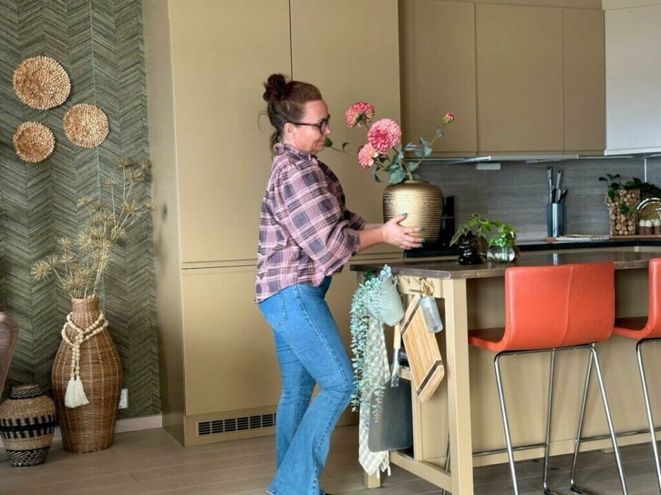 Sett prikken over i-en med dekor som fremhever de nye kjøkkenfrontene. (Foto: Lise Wolden) Lise Wolden, iført rutete skjorte og jeans, setter en gullvase med rosa blomster på kjøkkenøya. De nyoppussede, sandfargede kjøkkenfrontene gir rommet en helhetlig og innbydende stil.