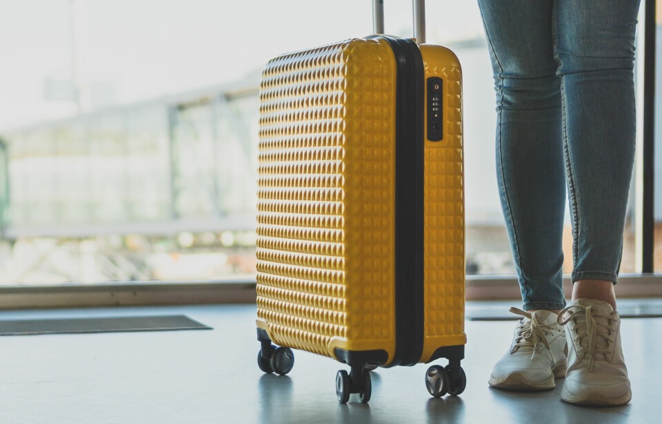 Stor koffert, Disse er best i test i 2025 En person står innendørs ved et stort vindu, sannsynligvis på en flyplass. Ved siden av dem står en knallgul koffert med hardt skall, strukturert overflate og fire hjul. Kofferten har innebygd kodelås, og håndtaket er trukket ut. Personen har på seg trange blå jeans og hvite joggesko. I bakgrunnen sees en glassgang eller jetbro som fører til et fly.