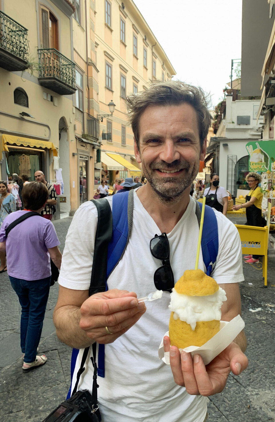 Gjermund Glesnes med en sitronsorbet servert i Amalfi-sitron under et besøk på den italienske Amalfikysten. «10 euro for en is er latterlig, men god, det var den», kommenterer Glesnes. Bildet viser en smilende mann som står i en sjarmerende gate omgitt av historiske bygninger og folkeliv – sannsynligvis et populært turistområde i Italia, mer presist Amalfikysten. Han holder en stor sitron med sitronsorbet og et lokk laget av skall, servert på lokal vis. Bak ham skimtes en bod med skiltet “Lemon Sorbet” og stabler av sitroner, noe som understreker stedets spesialitet. Amalfikysten lokker med idylliske småbyer og utsøkt mat, men for å få den ferien du drømmer om, må du vite hva du bør styre unna – og hvor de virkelige perlene ligger.