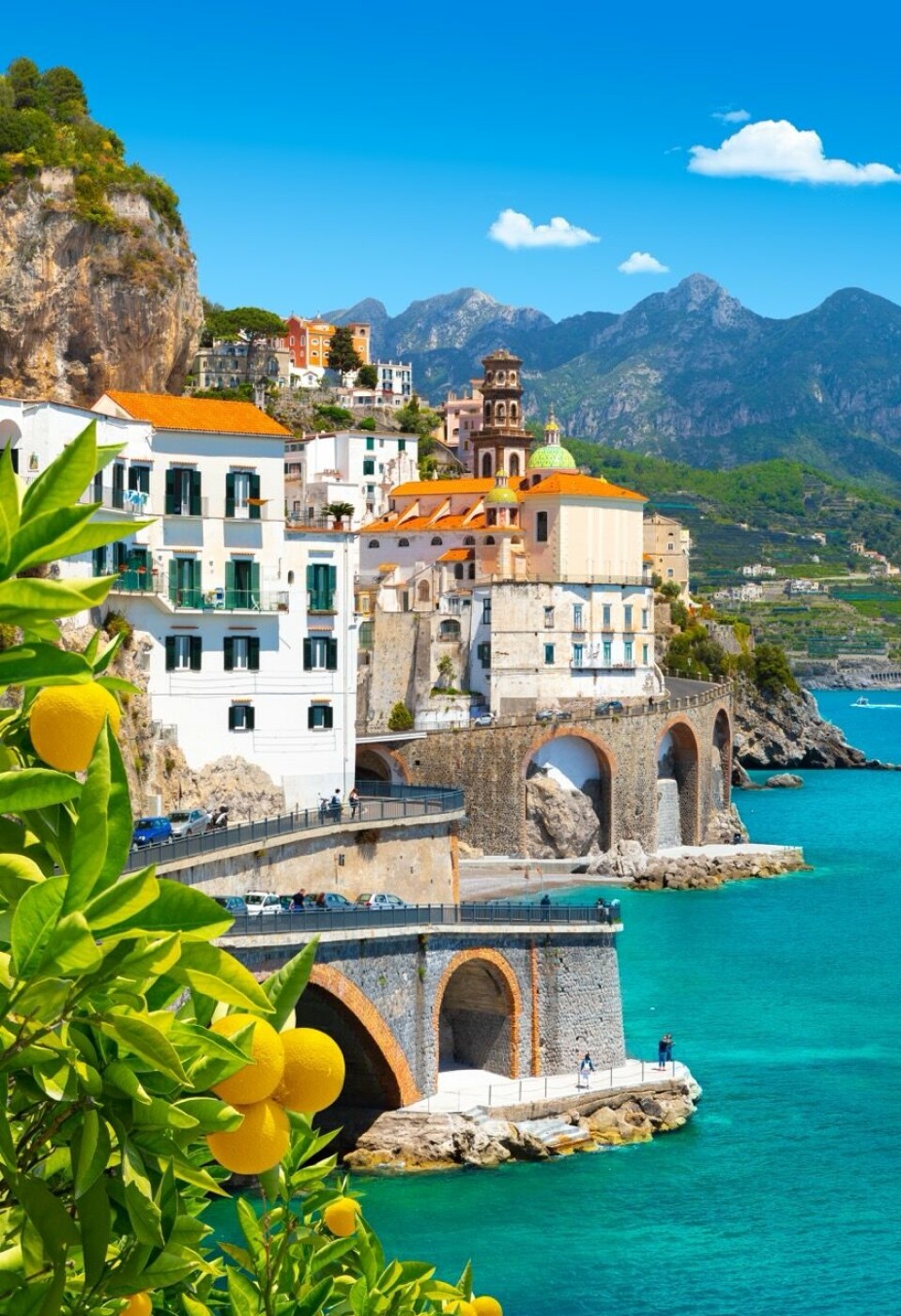 Vil du unngå de mest besøkte byene på Amalfikysten har Glesnes en klar anbefaling; – Det mest slående er Atrani, som ligger rett øst for Amalfi. Dette bildet viser nok en strålende utsikt over Atrani, en av Amalfikystens minste, men mest maleriske byer. I forgrunnen ser vi friske sitroner som symboliserer regionens berømte sitrondyrking. Den fargerike kirken med grønt og gult kuppeltak er Chiesa di San Salvatore de' Birecto, og de buede broene langs kystveien gir en dramatisk ramme rundt byen som stiger opp mot fjellene.