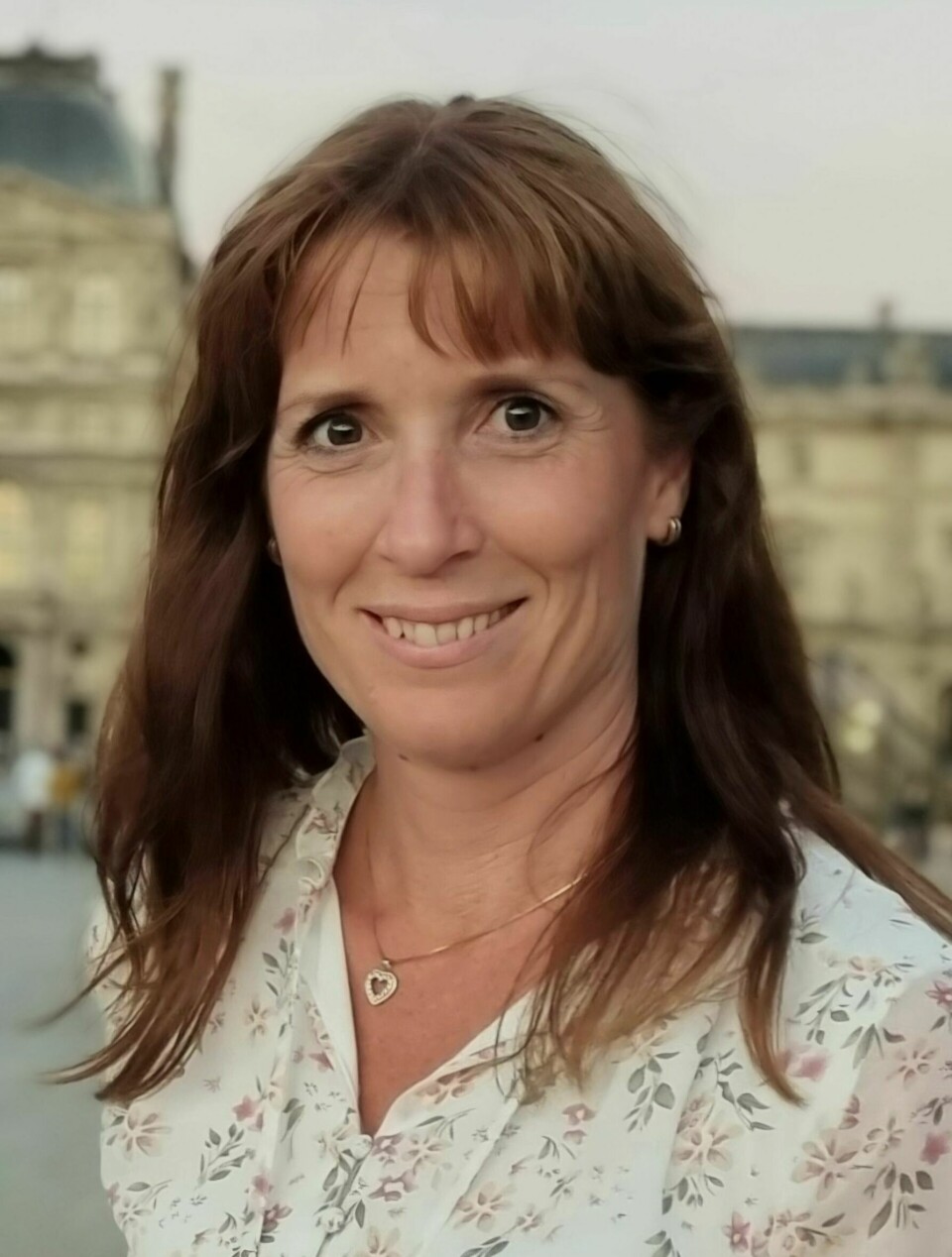 Cecilie Morland, professor ved Seksjon for farmakologi og farmasøytisk biovitenskap hos UiO. Bildet viser en kvinne som smiler vennlig til kamera. Hun har langt, brunt hår med pannelugg og bærer en lys bluse med blomsterprint. Hun har på seg en enkel halskjede med et hjerteanheng og små øreringer. I bakgrunnen skimtes en historisk bygning med klassisk arkitektur og kuppel, noe som antyder at bildet kan være tatt i et europeisk bymiljø, muligens Paris. Kvinnen fremstår rolig og profesjonell, og bildet er godt opplyst med et varmt kveldslys.
