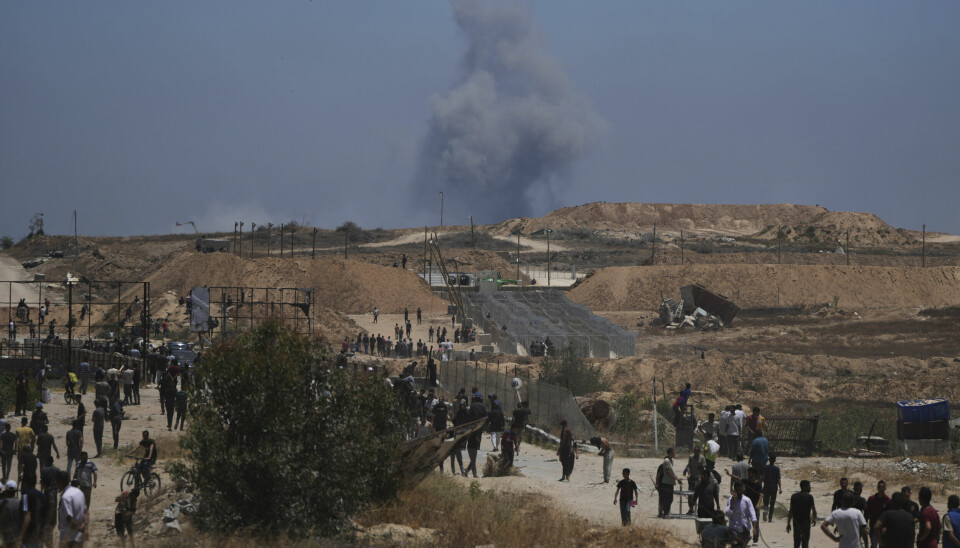 Palestinere ved en av nødhjelpsstasjonene til organisasjonen GHF i Khan Younis i slutten av mai. I bakgrunnen stiger røyk opp fra et israelsk angrep.
