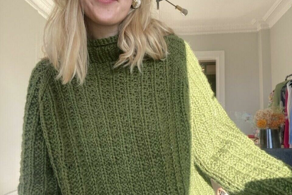 Skogdrøm-genseren fra Vild Knitwear – en tidløs, chunky strikk i dyp grønn nyanse. Perfekt for kalde dager og avslappet eleganse. En ung kvinne iført en chunky, strikket genser i en dyp skoggrønn farge. Genseren, kalt Skogdrøm, er fra Vild Knitwear og har en myk, ribbestrikket tekstur med en høy hals og vide ermer. Naturlig dagslys lyser opp plagget, og en koselig innendørs atmosfære skaper en varm og stilfull setting.