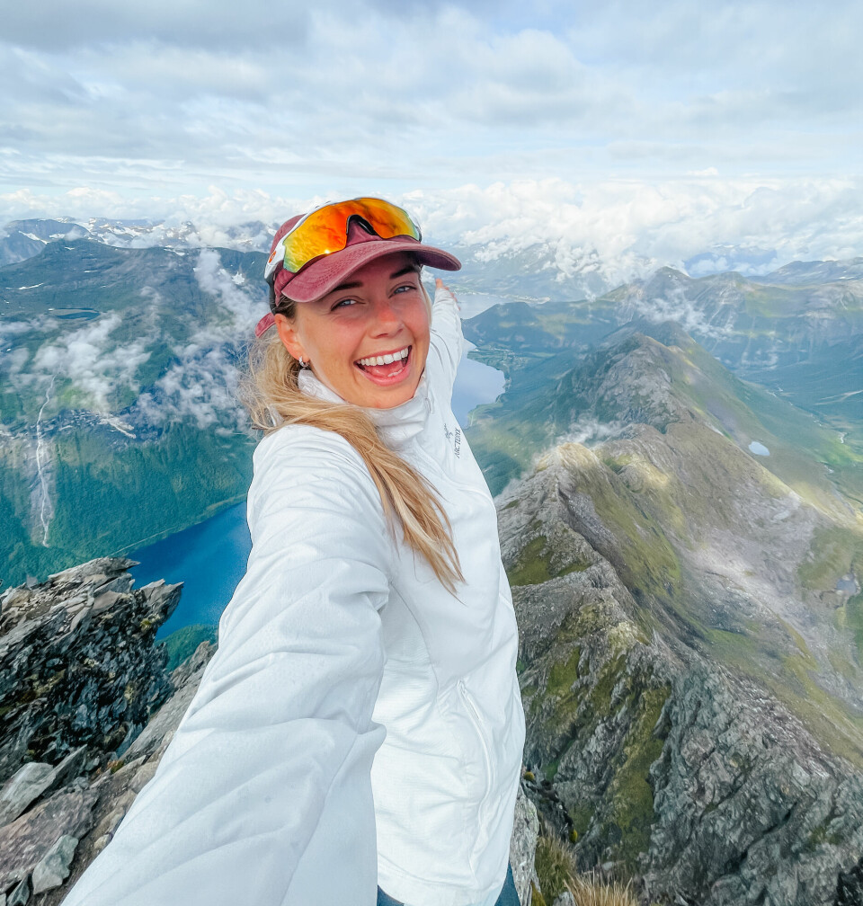 Veronica Solbak, redaktør for Ut.no, avslører årets mest populære fot- og fjellturer basert på søk på nettsidene deres. En smilende kvinne tar en selfie på toppen av et fjell med spektakulær utsikt i bakgrunnen. Hun har på seg hvit turjakke, rød caps og solbriller med speilglass som er løftet opp på hodet. I bakgrunnen ser man bratte fjellrygger, blå innsjøer og et teppe av skyer som henger lavt over landskapet – en dramatisk og imponerende utsikt som vitner om norsk høyfjell på sitt mest majestetiske. Stemningen i bildet er energisk og glad, og det tydeliggjør gleden ved å nå toppen etter en krevende fjelltur.