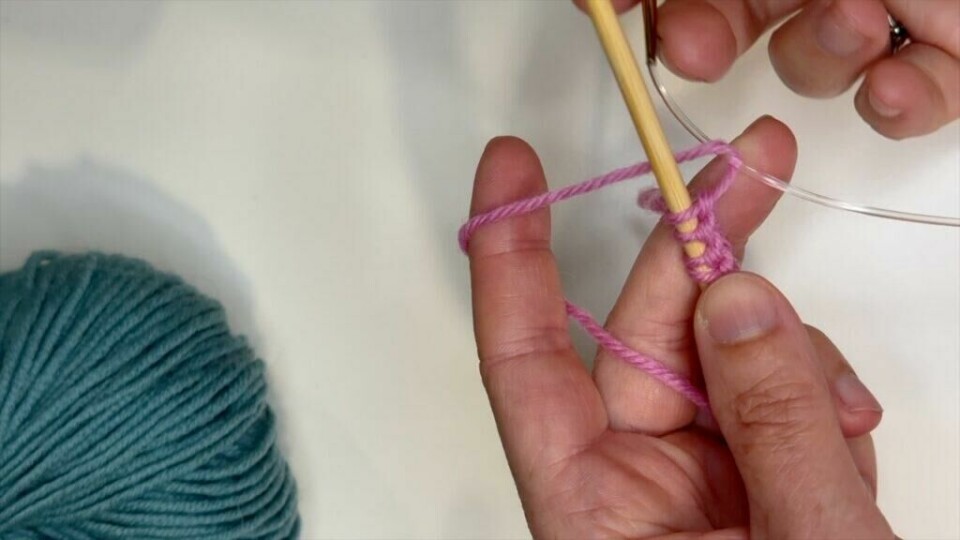 Se hvordan du fanger inn rundpinnen med garnet du strikker med. (Foto: Kreative Ideer/ Miriam Schjetlein) Hender som holder et strikketøy med rosa garn. Det vises hvordan man kan plukke opp masker underveis i strikkingen med en ekstra rundpinne.