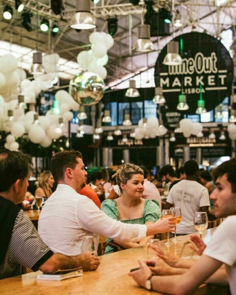 Time Out Market i Lisboa er en populær innendørs mathall som tilbyr et bredt utvalg av portugisisk mat. Bildet viser et matmarked, spesifikt Time Out Market i Lisboa. Folk sitter ved lange trebord og nyter mat og drikke. En mann i en hvit skjorte og en kvinne i en grønn kjole sitter sammen og skåler med glassene sine. Rundt dem er det andre besøkende som også nyter måltidene sine. Taket er dekorert med hvite ballonger og flere hengende lamper, og en stor diskokule henger i midten, som reflekterer lysene i rommet. Atmosfæren virker livlig og sosial, med mange mennesker samlet for å nyte markedets tilbud. Her finner du tips til ni matmarkeder i populære europeiske storbyer som Paris, Berlin, Roma, Barcelona, København, Lisboa og London.