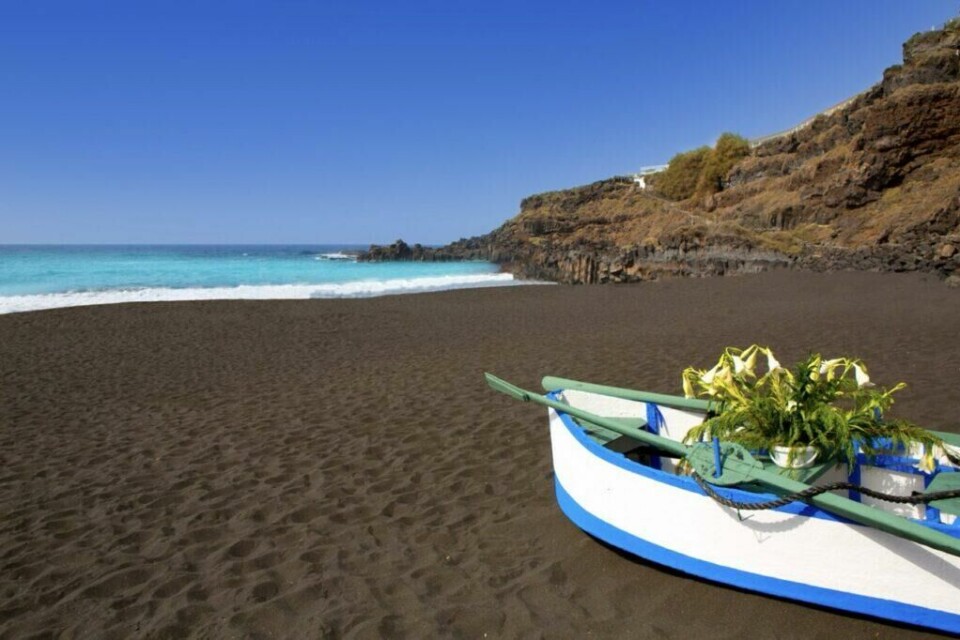 Vil du utforske vulkanske strender med mørk sand, anbefales det å ta turen til avbildede El Bollullo nord på Tenerife. (Foto: Canva) Bildet viser en idyllisk strand med svart vulkansk sand, typisk for Tenerifes unike landskap. I forgrunnen står en liten, malt trebåt i hvitt og blått, dekorert med en vase fylt med frodige grønne planter og blomster. Havet i bakgrunnen er krystallklart og glitrende turkis, med hvite bølger som bryter mot stranden. Kystlinjen er omgitt av bratte, steinete klipper som tilfører dramatikk til det rolige strandbildet. Den klare, blå himmelen setter prikken over i-en og fremhever det naturskjønne miljøet som Tenerife er kjent for. Kjent for sine vakre og unike strender, et storslagent karneval og Europas mest besøkte park. Dette vil du ikke gå glipp av på kanariøya Tenerife!