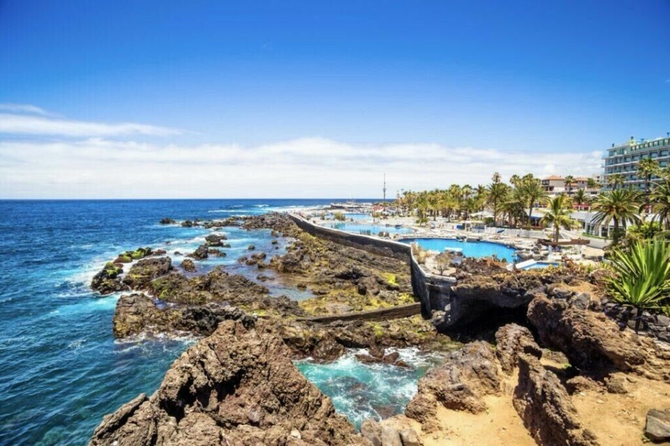 Bilde av Lago Martiánez i Puerto de la Cruz, Tenerife. Et unikt bassengkompleks på den største av Kanariøyene designet av kunstneren César Manrique. (Foto: Canva) Bildet viser Lago Martiánez i Puerto de la Cruz, Tenerifes, et vakkert kystlandskap med mørke vulkanske klipper som strekker seg ut i det turkise havet. På høyre side er det et stort, velorganisert rekreasjonsområde med svømmebassenger omgitt av palmer, solsenger og bygninger, sannsynligvis en del av en populær turistattraksjon. Himmelens klare blåfarge og solskinnet skaper en idyllisk stemning. Dette ser ut til å være et tropisk reisemål, perfekt for avslapning og nytelse av naturen.