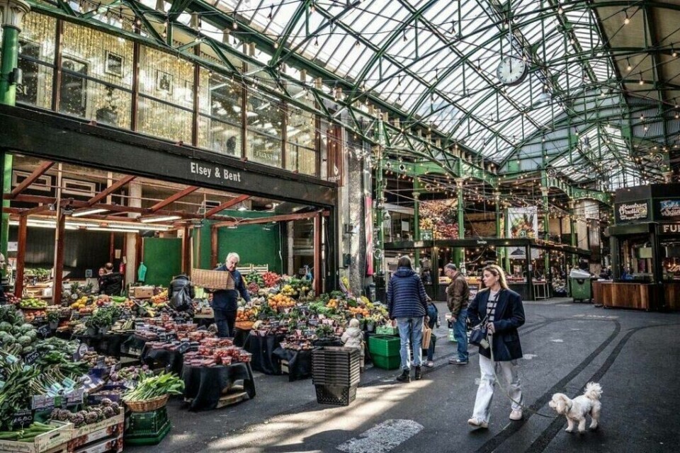 Borough Market er et av de eldste og største matmarkedene i London. (Foto: Borough Market) Bildet viser et travelt innendørs matmarked i London, nærmere bestemt Borough Market. Flere mennesker går rundt og handler frukt og grønnsaker fra forskjellige boder. En eldre mann bærer en kasse med varer gjennom markedet. Markedet har et høyt tak med grønne metallbjelker og glasspaneler som slipper inn naturlig lys. Lyslenker og dekorative lamper henger ned fra taket, og gir en varm og innbydende atmosfære. Her finner du tips til ni matmarkeder i populære europeiske storbyer som Paris, Berlin, Roma, Barcelona, København, Lisboa og London.