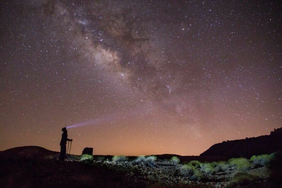 Tenerife er en av verdens beste steder for stjernekikking og på kanariøya befinner også verdens største solobservatorium seg. (Foto: Canva) Bildet viser en klar stjernehimmel med Melkeveien som hovedfokus, strålende på tvers av himmelen i en myriade av lys og farger. Forgrunnen består av et øde landskap med sparsomme planter og en silhuett av en person som står med en lyskaster, rettet mot stjernene. Lyset fra lyskasteren skaper en dramatisk effekt som trekker blikket mot himmelen. Horisonten har et varmt, gyllent skjær, sannsynligvis fra en solnedgang eller lysforurensning fra fjerne byer. Bildet fanger en følelse av undring og ro i møte med universets storhet, perfekt for stjernetitting og astrofotografi. Kjent for sine vakre og unike strender, et storslagent karneval og Europas mest besøkte park. Dette vil du ikke gå glipp av på kanariøya Tenerife!