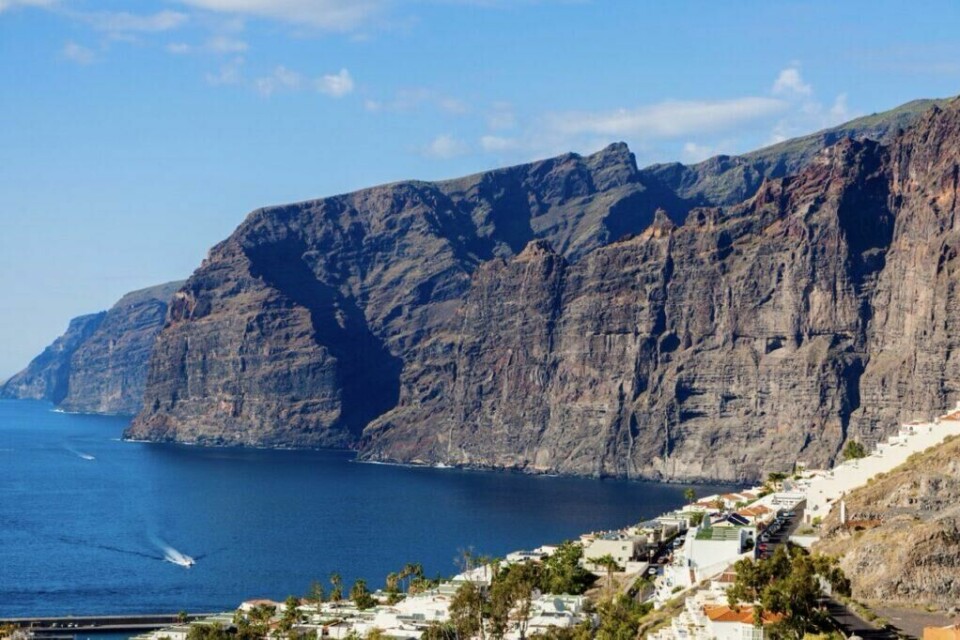 Klippene Acantilados de Los Gigantes, som på norsk betyr «kje,pene», stiger opp fra havet til en høyde på 500-800 meter. (Foto: Canva) Bildet viser de imponerende klippene Acantilados de Los Gigantes, som strekker seg dramatisk opp fra havet langs Tenerifes vestkyst. Disse massive klippeformasjonene, som kan nå høyder på opptil 600 meter, dominerer landskapet og skaper en dramatisk kontrast til det dype blå havet. I forgrunnen ser vi en kystby med hvitkalkede bygninger som klatrer oppover terrenget, omgitt av frodig vegetasjon. En liten båt er synlig på vannet, og den klare blå himmelen bidrar til den spektakulære utsikten. Dette stedet er kjent som et populært reisemål for både naturopplevelser og båtturer. Kjent for sine vakre og unike strender, et storslagent karneval og Europas mest besøkte park. Dette vil du ikke gå glipp av på kanariøya Tenerife!