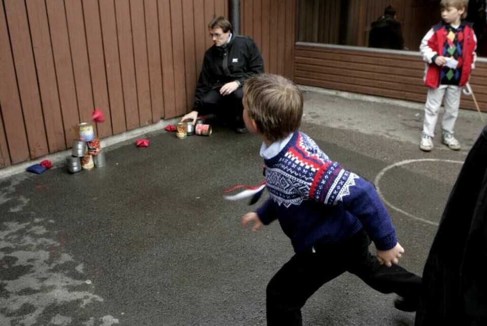 Llek og moro på Hallagerbakken skole i Oslo. Regnvær preget feiringen av nasjonaldagen, men ball- eller sandpose-kast på bokser er like morsomt uanset vær.Foto Cornelius Poppe / NTB (Foto: Cornelius Poppe / NTB) På bildet ser vi en gutt i mørk bukse og blå strikkegenser med tradisjonelt mønster kaste en liten pose mot en stabel med tomme hermetikkbokser. En voksen mann sitter på huk ved stabelen og følger med, klar til å stable boksene på nytt. I bakgrunnen ser vi flere barn, hvorav ett holder en pinne og observerer leken. Bildet viser den klassiske 17. mai-leken «boksekast», der deltakerne prøver å velte flest mulig bokser med en ertpose. Underlaget er vått asfalt, og omgivelsene gir inntrykk av at leken foregår i skolegård eller utenfor et forsamlingslokale. Dette er en tradisjonell og lavterskel lek som ofte brukes i 17. mai-feiring for barn.