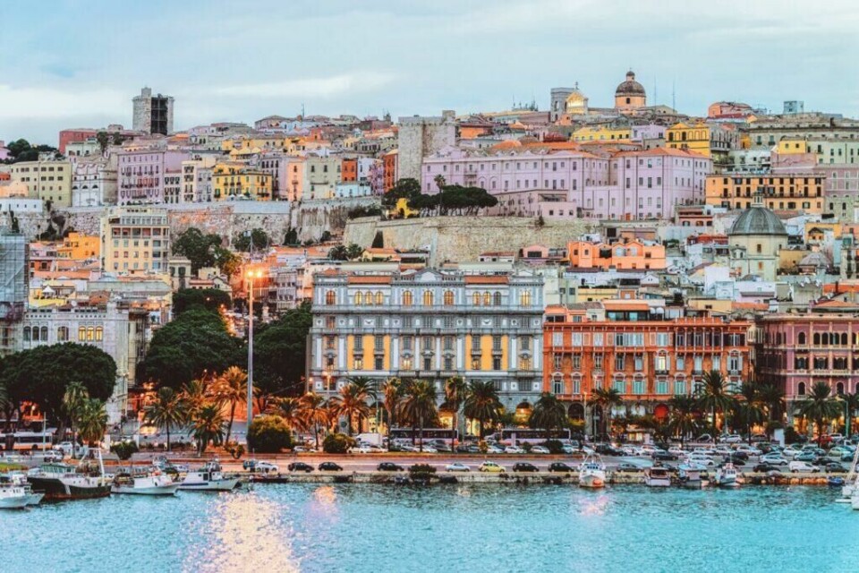 Hovedstaden på Sardinia, Cagliari, er også verdt å besøke. Her finner du sjarmerende gater og pastellfargede hus. (Foto: Colourbox) Et fargerikt bilde av byen Cagliari på Sardinia, Italia. Bildet viser en rekke bygninger i pastellfarger som ligger tett sammen på en høyde, med noen eldre strukturer og tårn som stikker opp. Ved foten av bakken er det en rekke palmer og en travel havnepromenade med parkerte biler og båter fortøyd ved kai. Lyset fra gatelykter og vinduer gir en varm glød. Middelhavsøya Sardinia byr på både luksus og spennende historie, og her finner du krystallklar sjø og kritthvite strender. Vi gir deg tips til hva du kan se og gjøre på den italienske øya i Middelhavet; Dra til Smaragdkysten, Grotta di Nettuno, Nuraghe-ruinene, La Maddalena-øygruppen. Vi hjelper også med å finne hotell på Sardinia.