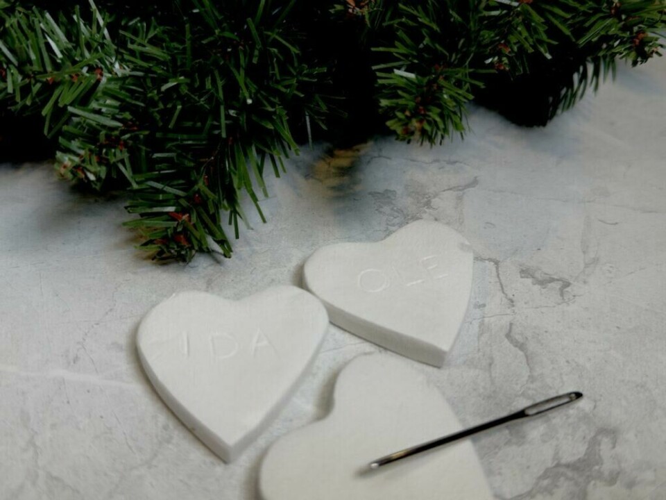 Personlig juletrepynt av leire – enkel og sjarmerende DIY for julegavene (Foto: Kreative Ideer/Aurora Lange) Hjerteformede juletrepynt i selvtørkende leire med navnene 'IDA' og 'OLE'. Lappene er plassert ved en grønn gren og en nål, snart klare for å henges opp i juletreet.
