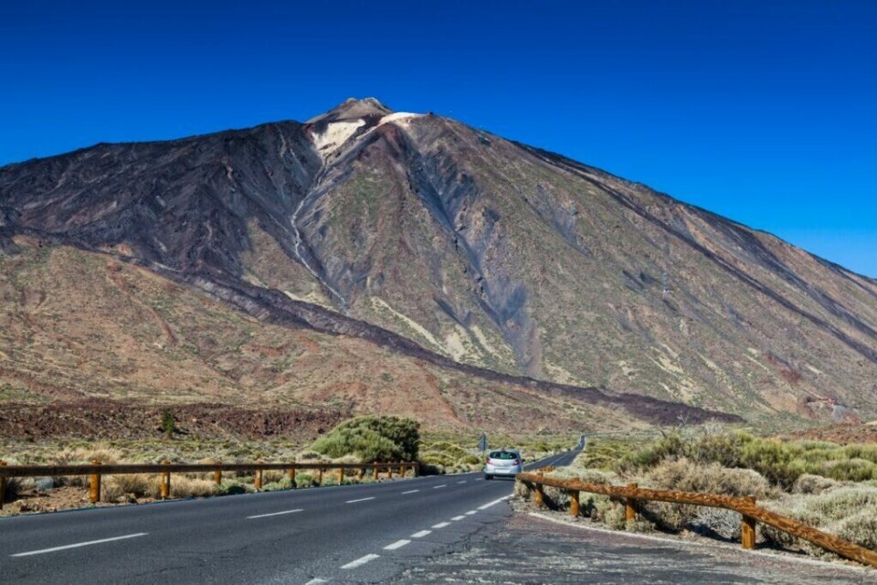 Spanias høyeste fjell, Teide, finner du på Tenerife. (Foto: Colourbox) En bil kjører langs en vei som leder mot fjellet Teide på Tenerife, Kanariøyene. Fjellet er dekket av mørke vulkanske steiner og vegetasjon, og den klare blå himmelen gir en skarp kontrast til landskapet. Veien er omgitt av busker og små trær. Tenerife er den største Kanariøyen og har Spanias høyeste fjell, Teide. Hovedstaden er Santa Cruz de Tenerife. Her kan du finne billige reiser til Tenerife, samt hotell på den populære kanariøyen.