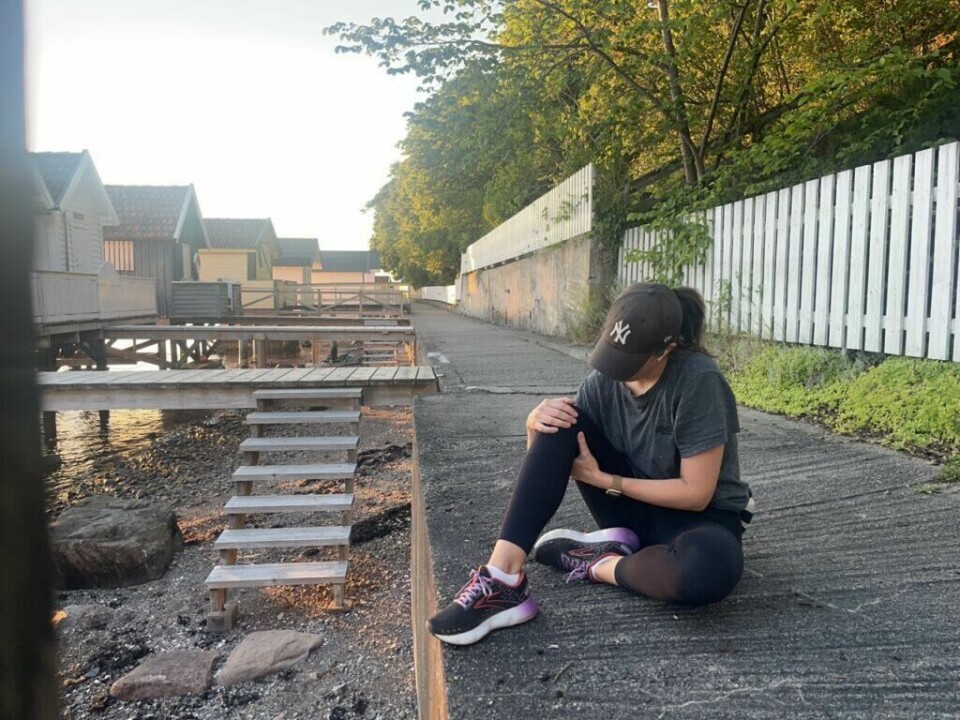 Bilde av en kvinne som sitter på en gangvei ved små badehus ved Oslofjorden. Hun har på seg en grå treningst-skjorte, en svart NY-Yankees caps og svarte tights. Hun holder på kneet sitt fordi hun kjenner smerter og vondter og har en smartklokke på håndleddet. Bakgrunnen viser små badehus og blå himmel ved Oslofjorden. Solen skinner, og det er en lys og fin dag. Løpeekspert Andreas Gossner fra Løpetrening.no forteller (Foto: Cecilie Aasland / ABC Startsiden)