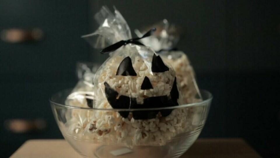 Popcorn-pakker med gresskar-ansikter, perfekt for halloween-snacks (Foto: Kreative Ideer/ Aurora Lange) Bildet viser gjennomsiktige poser fylt med popcorn, plassert i en glassbolle. Hver pose er dekorert med sorte gresskar-ansikter laget av papir, og er bundet med svarte bånd på toppen for å gi en ekstra halloween-stemning. Posene gir inntrykk av å være små gresskar og utgjør en kreativ og morsom snacksidé til halloween-festen. Belysningen er dempet, noe som gir bildet et koselig og litt mystisk preg.