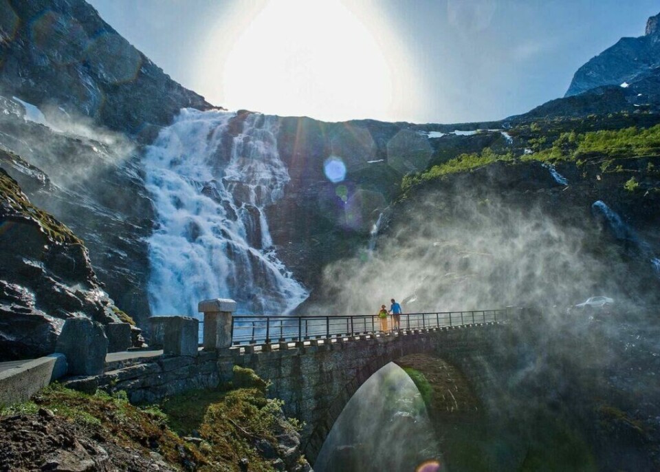 Geiranger og Trollstigen er virkelig en perle på Vestlandet du ikke vil gå glipp av. (Foto: Fjord Norway) En stor foss stuper nedover fjellsiden ved Trollstigen, mens solen skinner klart i bakgrunnen. Vannet spruter og lager en fin tåke som omgir området. En steinbro med rekkverk krysser fossen, og på broen står to personer som ser ut til å nyte synet av fossen. Fjellene rundt er dekket med noe vegetasjon og snøflekker. Her får du tips til syv vakre bilturer i Norge som er perfekt for norgesferien. Geiranger og Trollfjorden, Atlanterhavsveien, Lofoten, Hardanger, Rondane, Gamle Strynefjellsvegen, Senja.