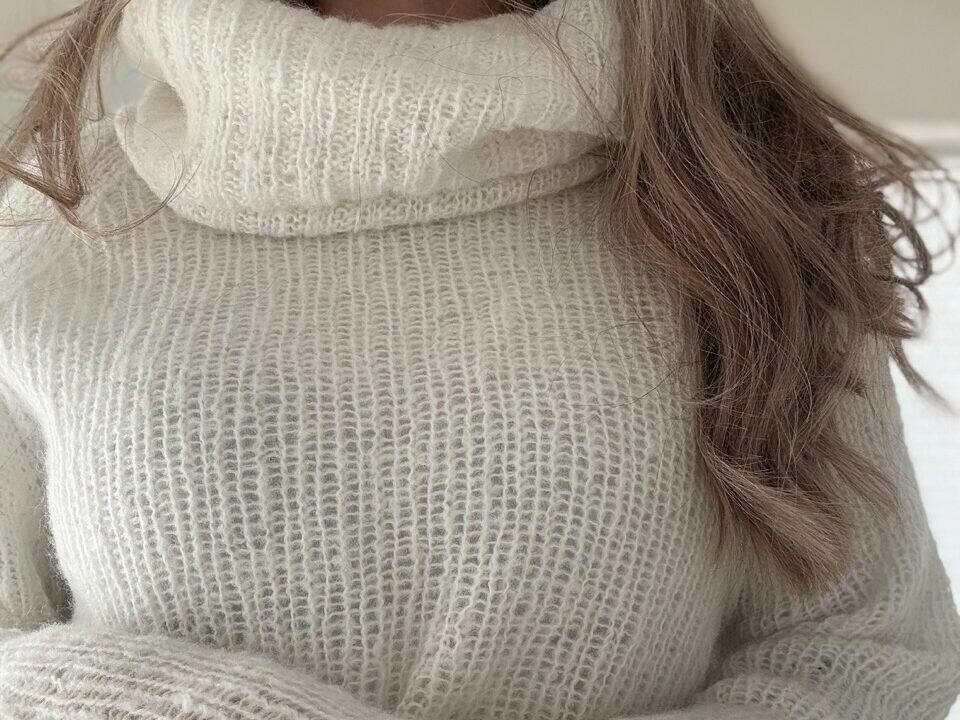 Agneta-genseren sett forfra. Genseren er designet av Ull og Strikk. Agneta-genseren i en avslappet styling, kombinert med lyse jeans. Den hvite ribbestrikkede genseren har en høy hals som kan brettes ned for ekstra komfort. Mykt og luftig materiale gjør den ideell for kjølige dager, samtidig som den gir et moderne, minimalistisk uttrykk.