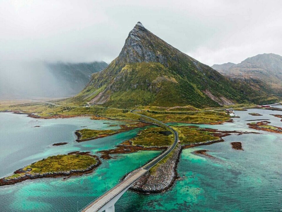 Hvem drømmer vel ikke om å oppleve vakre Lofoten, hvor hvite strender og krystallklart hav møter spisse fjell. (Foto: Joshua Kettle/Unsplash) Et luftfoto som viser en lang bro i Lofoten som strekker seg over klart, turkisfarget vann og forbinder små øyer. Den spisse toppen av Volandstinden stiger majestetisk opp fra det grønne landskapet, omgitt av lavtliggende skyer og tåkedekte fjell i bakgrunnen. Her får du tips til syv vakre bilturer i Norge som er perfekt for norgesferien. Geiranger og Trollfjorden, Atlanterhavsveien, Lofoten, Hardanger, Rondane, Gamle Strynefjellsvegen, Senja.
