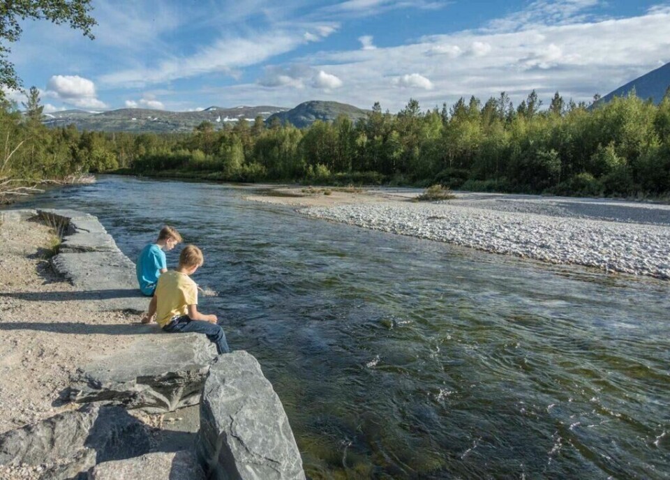 Rondane er et fjellområde i Midt-Norge som er kjent for sin ville og uberørte natur. (Foto: Tonje Tjernet/Statens vegvesen) To barn sitter på en steinkant ved siden av en elv i Rondane. De ser ut til å leke med vannet, og en av dem har føttene i elven. Landskapet rundt elven er frodig med trær, og i bakgrunnen kan man se fjell med snøflekker under en klar, blå himmel med spredte skyer. Elven flyter rolig gjennom det naturskjønne området. Her får du tips til syv vakre bilturer i Norge som er perfekt for norgesferien. Geiranger og Trollfjorden, Atlanterhavsveien, Lofoten, Hardanger, Rondane, Gamle Strynefjellsvegen, Senja.