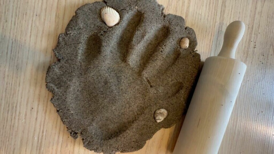 Bruk sandleire til å lage hånd- eller fotavtrykk. Pynt med skjell og sneglehus. (Foto: Kreative Ideer/ Miriam Schjetlein) Bildet viser en flatt utkjevlet sirkelformet del av sandleire som ligger på et lyst trebord. I leiren er det laget et tydelig håndavtrykk. Rundt håndavtrykket er det dekorert med tre små skjell som er presset ned i leiren. Ved siden av leiren ligger en kjevle i tre, som har blitt brukt til å rulle ut leiren til ønsket tykkelse. Dette er en kreativ aktivitet, som involverer å lage personlige minner med sandleire ved hjelp av håndavtrykk og naturlige dekorasjoner som skjell.