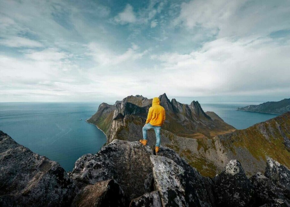 Opplev vakre Senja i Troms. Her får du alt fra spektakulære fjell til vakre sandstrender. (Foto: Ansgar Scheffold/Unsplash) En person iført en gul jakke og jeans står på en fjellknaus og ser utover det dramatiske landskapet på Senja. Foran personen strekker fjellene seg ut mot havet med bratte klipper og spisse topper. Under ligger det blå havet, og horisonten er dekket av en lett skyet himmel. Utsikten gir et imponerende og majestetisk inntrykk av naturen. Her får du tips til syv vakre bilturer i Norge som er perfekt for norgesferien. Geiranger og Trollfjorden, Atlanterhavsveien, Lofoten, Hardanger, Rondane, Gamle Strynefjellsvegen, Senja.