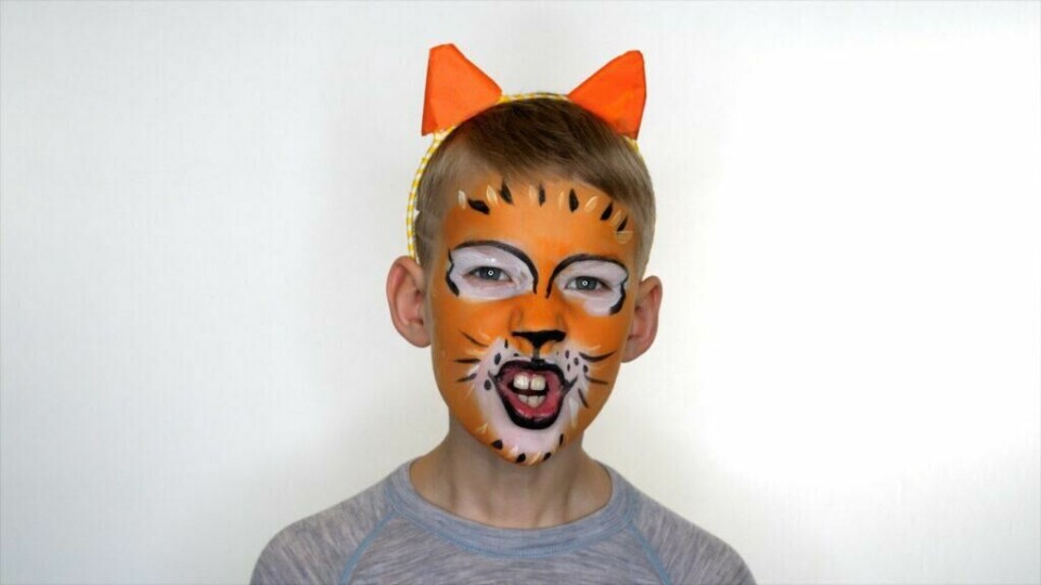 Slipp løs din indre tiger med fargerik ansiktsmaling! (Foto: Kreative Ideer/ Miriam Schjetlein) Bildet viser et barn med et kreativt tigeransiktsmalingdesign. Ansiktet er malt i klare oransje toner, med svarte striper og hvite detaljer rundt øynene og munnen for å etterligne en tiger. Barnet har på seg et matchende pannebånd med oransje ører, som forsterker tigerlooken. Han har en entusiastisk uttrykk, med åpen munn, noe som gir liv til det ville dyret han etterligner. Bakgrunnen er nøytral, og barnet har på seg en enkel grå genser.