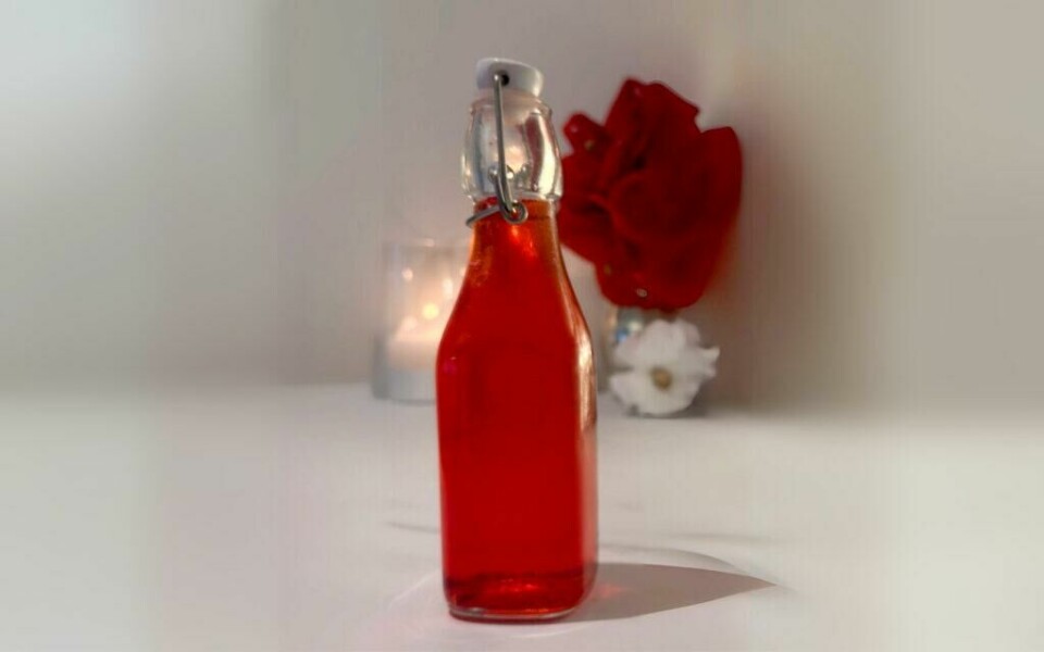 Hjemmelaget jordbærvinaigrette i dekorativ flaske – en smakfull og personlig julegave laget av jordbærsirup og eddik. Et nærbilde av en flaske fylt med hjemmelaget jordbærvinaigrette. Den røde vinaigretten står i en dekorativ glassflaske med patentkork, omgitt av enkel bakgrunnsdekor som levende lys og blomster. Perfekt som julegave, laget av hjemmelaget jordbærsirup og hvitvinseddik.