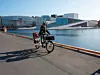Bilde av en dame på en svart laste-elsykkel med en svart sykkelhjelm, en svart boblejakke og utsikt til Oslofjorden, Operaen og Bjørvika. Damen har et barn fremme på el-lastesykkelen sin. Hvem en elektrisk lastesykkel passer til, hvilken type du bør velge og hvordan du velger riktig til ditt bruk fikk vi alle svarene på da vi forhørte oss med Janne Pedersen fra NAF. Hun forteller også hva en longtail-sykkel er, hva du bør velge av to eller tre hjul på en el-lastesykkel, hvordan du velger lastesykkel om du skal frakte med deg barn og hva som er forskjellen på en billig lastesykkel og en dyr. Dette håper vi gjør det enklere å velge den beste elektriske lastesykkelen for deg!