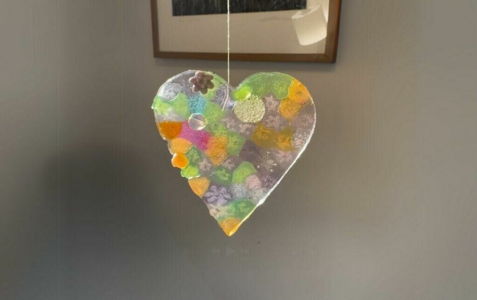 Heng fargerik glede i vinduet med hjemmelagde glassmalerier av plastperler. (Foto: Kreative Ideer/ Miriam Schjetlein) Et vakkert hjerteformet glassmaleri laget av smeltede plastperler henger i en tynn tråd foran en lys bakgrunn. Hjertet har klare, livlige farger som oransje, grønn, gul, rosa og lilla, og gir et mosaikkaktig preg. Lyset fra vinduet bak fremhever den glitrende, gjennomsiktige effekten av perlene. I bakgrunnen skimtes en innrammet veggdekorasjon, noe som gir en lun og hjemlig atmosfære.
