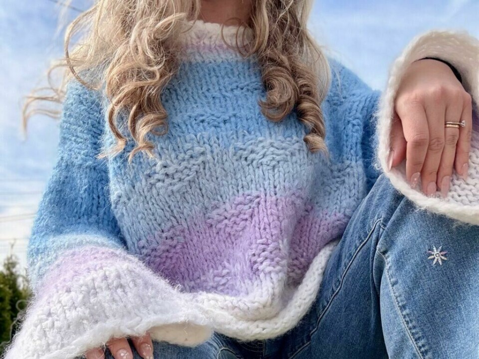 Bryggegenseren strikkes med dobbel tråd i fnuggmykt garn og har en fargepalett som minner om solnedgang ved sjøen. (Foto: Kreative Ideer/ Aurora Lange) Nærbilde av Bryggegenseren i bruk, strikket i Camilla Pihl Fnugg med myk fargeskalering fra hvit til lavendel og himmelblå. Genseren har et strukturert hjertemønster, vide ermer og myke overganger mellom fargene.