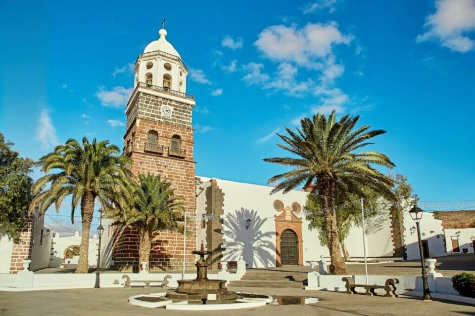 Besøker du Lanzarote kan vi anbefale å ta turen til torget i byen Teguise. Kanariøya har også mange andre spennende ting som er verdt å få med seg. (Foto: Colourbox) Et historisk klokketårn omgitt av palmetrær i en pittoresk by på Lanzarote, Kanariøyene. Klokketårnet står på en liten plass med en fontene i forgrunnen, omgitt av hvitmalte bygninger med tradisjonell arkitektur under en lys blå himmel.