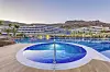 Bildet viser det elegante Radisson Blu Resort & Spa, Gran Canaria, omgitt av fjell og tropiske palmer. I forgrunnen ser vi et boblebad med blått mosaikkflis, som leder videre til et stort, krystallklart basseng. Hotellets moderne bygning strekker seg i en halvmåneform, med romslige balkonger som gir utsikt mot bassenget og de vakre omgivelsene. Atmosfæren er avslappende og luksuriøs, perfekt for en ferie på Gran Canaria.