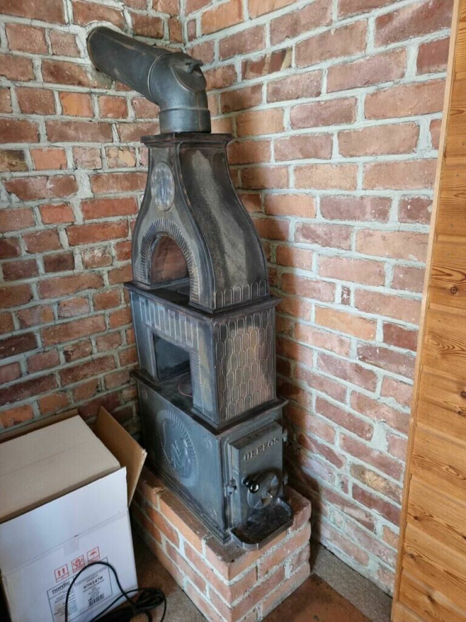 Flere kommuner har som et tiltak innført panteordninger på gamle vedovner og dette gjelder blant annet Oslo og Bergen. Bildet viser en eldre, dekorativ vedovn i støpejern plassert mot en vegg av rød teglstein. Ovnen har en rustikk stil med detaljerte mønstre og ornamenter, og den er montert på en sokkel av murstein. Et rør for røykgass går fra toppen av ovnen og inn i veggen. Til venstre for ovnen står en pappeske med synlige etiketter og en elektrisk ledning, som gir en kontrast til den gamle ovnens tradisjonelle stil. På høyre side kan man se en del av en vegg med lyst treverk, som gir rommet et varmt og naturlig preg. Ovnen ser ut til å være i god stand, men den representerer en eldre type som kan være omfattet av moderne restriksjoner for bruk eller installasjon. Har du kjøpt en brukt vedovn? Da kan det være ulovlig å installere den! Vi har snakket med eksperten som forteller hvilke det gjelder.