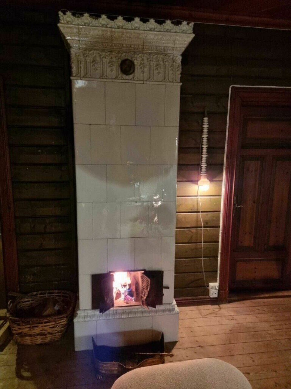 Regelen om ulovlig installasjon av gamle vedovner har noen viktige unntak og det er blant annet at alle klebersteins- og kakkelovner kan flyttes å brukes videre. (Foto: Jan-Loek Siskens (ABC Startsiden)) Bildet viser en klassisk, hvit kakkelovn i et rom med mørke trevegger og tregulv. Ovnen har et dekorativt mønster øverst, og døren til ildstedet står åpen, med synlige flammer som skaper en varm og koselig stemning. Til venstre for ovnen står en flettet vedkurv fylt med ved, og foran ovnen står en metallbeholder for aske eller tilbehør. På høyre side av bildet henger en enkel lyspære fra en ledning på veggen, som gir et svakt, varmt lys til rommet. Ved siden av ovnen er en dør av tre med tydelige årringer og detaljer, som understreker det tradisjonelle og rustikke interiøret.Har du kjøpt en brukt vedovn? Da kan det være ulovlig å installere den! Vi har snakket med eksperten som forteller hvilke det gjelder.