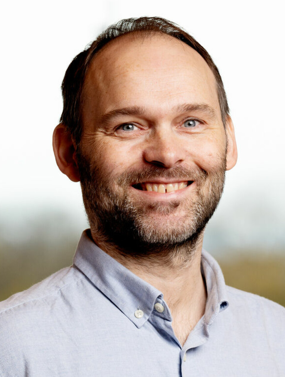 Craig Bøthun jobber hos SpareBank 1 Sør-Norge. En smilende mann i lyseblå skjorte står foran et vindu med uskarp bakgrunn av høstfargede trær. Han har kort, mørkt hår, skjegg og ser direkte mot kamera med et åpent og vennlig uttrykk.