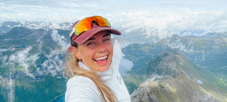 En smilende kvinne tar en selfie med én arm strukket ut over kanten av et stup. Bak henne strekker et dramatisk fjellandskap seg ut, med dype fjorder, grønne dalsøkk og skybanker som driver over tindene. Bildet er fullt av energi og turglede, og fanger følelsen av mestring og frihet etter en luftig topptur – et høydepunkt for mange som går Slogenrunda.