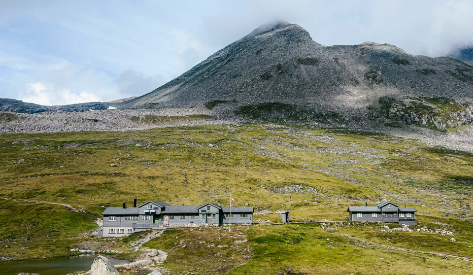 Midt i et nakent og vidstrakt fjellandskap ligger en stor DNT-hytte omgitt av lav, grønn vegetasjon og små dammer. Bak hytta reiser et grått fjellmassiv seg med en tåkedott på toppen. Dette er Patchellhytta, en sentral base for fjellturer i Sunnmørsalpene, og et naturlig stoppested på den populære rundturen Slogenrunda. Her kan man hvile etter krevende stigninger og nyte utsikten mot noen av Norges mest ikoniske fjell.
