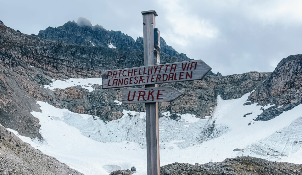Et veivisningsskilt står blant stein og snø, omgitt av alpine fjell. Skiltet peker én vei mot «Patchellhytta via Langesæterdalen», og den andre mot «Urke». Dette er et viktig knutepunkt i fjellheimen på Sunnmøre, hvor mange starter eller avslutter Slogenrunda – en rundtur som tar deg gjennom frodige daler, isbrelandskap og ikoniske topper i Sunnmørsalpene.