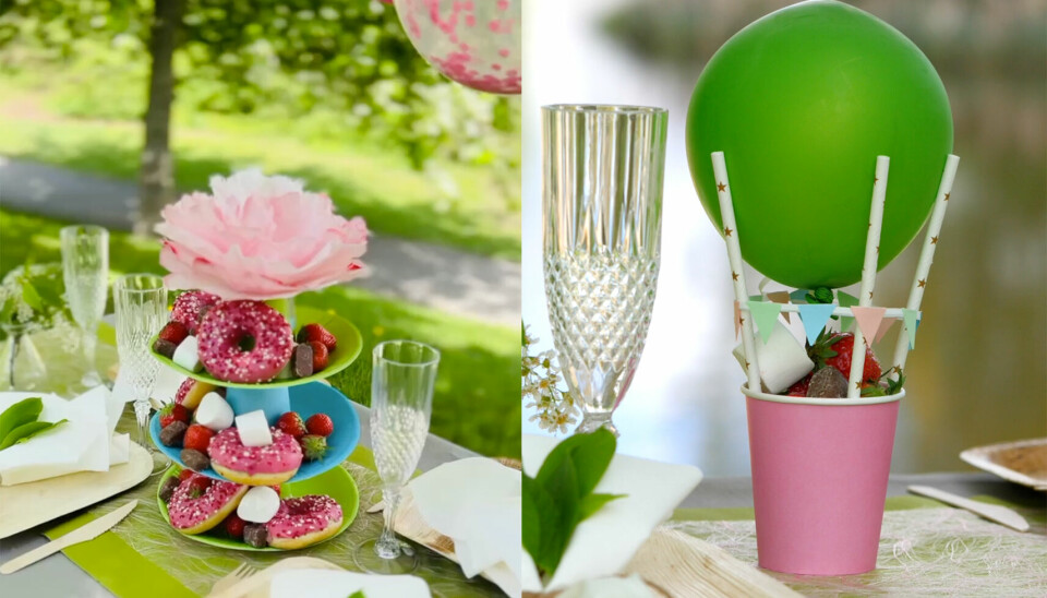 Festbord med hjemmelaget etasjefat og luftballong-godteskål laget av enkle materialer som papp og sugerør. Bildet viser to DIY-ideer for borddekking til fest: Til venstre et fargerikt etasjefat laget av papptallerkener og kopper, pyntet med en stor rosa papirblomst og fylt med donuts, jordbær og marshmallows. Til høyre en godteskål utformet som en luftballong, laget med en rosa pappkopp, grønne sugerør, en grønn ballong og små girlandere, fylt med jordbær og godteri. Bordet er dekket med naturinspirerte detaljer og glass med mønster.