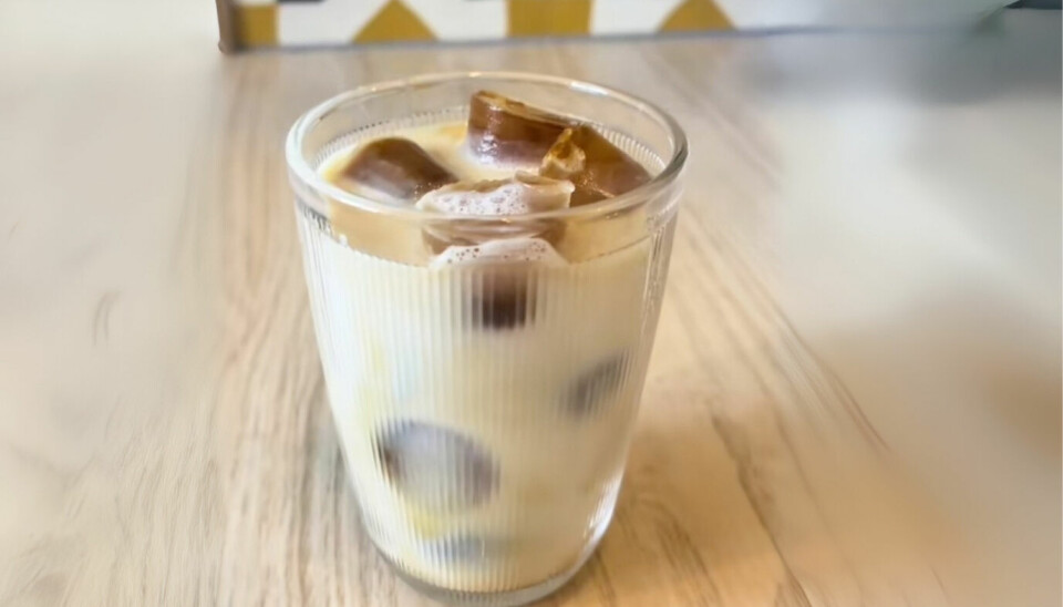 Lag iskaffe med kaffeisbiter for en fyldig smak uten utvanning. Et glass med iskaffe laget med kaffeisbiter og melk står på et lyst trebord. De mørke kaffeisbitene flyter i melken, som gir et delikat lagdelt uttrykk. Bakgrunnen viser et moderne flisemønster i sort, hvitt og gult.